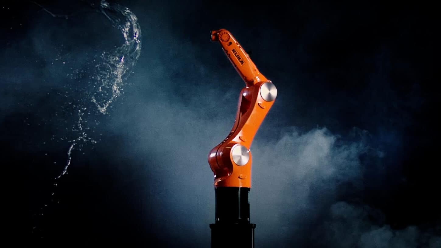 Der orangefarbene KUKA Agilus Roboterarm wird, vor einem dunklen, nebeligem Hintergrund, mit Wasser bespritzt.