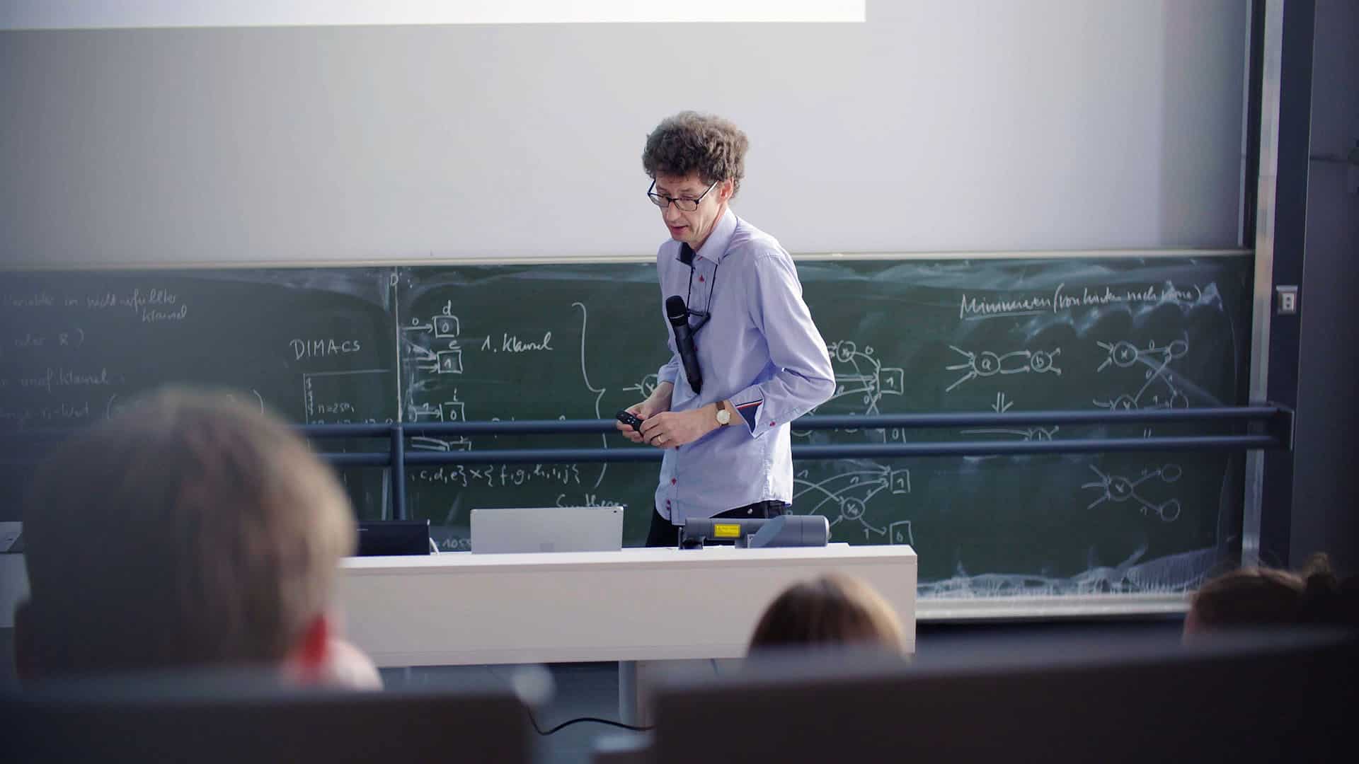 Ein Dozent der Uni Ulm hält mit einem Mikrofon um den Hals hängend eine Vorlesung vor vollem Hörsaal.